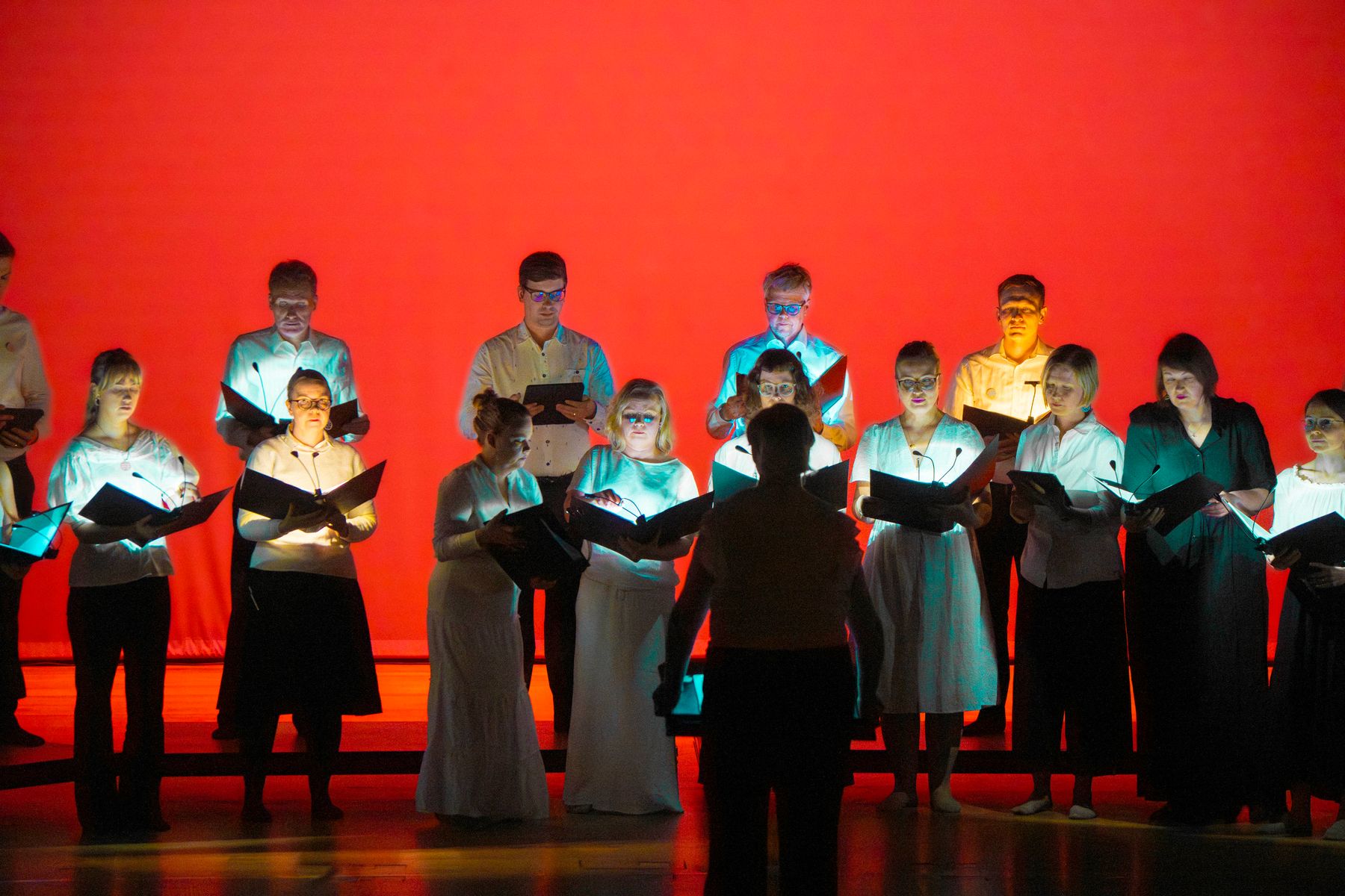 Kuorolaisia ja johtaja esiintymässä kirkkaanpunaisessa valaistuksessa