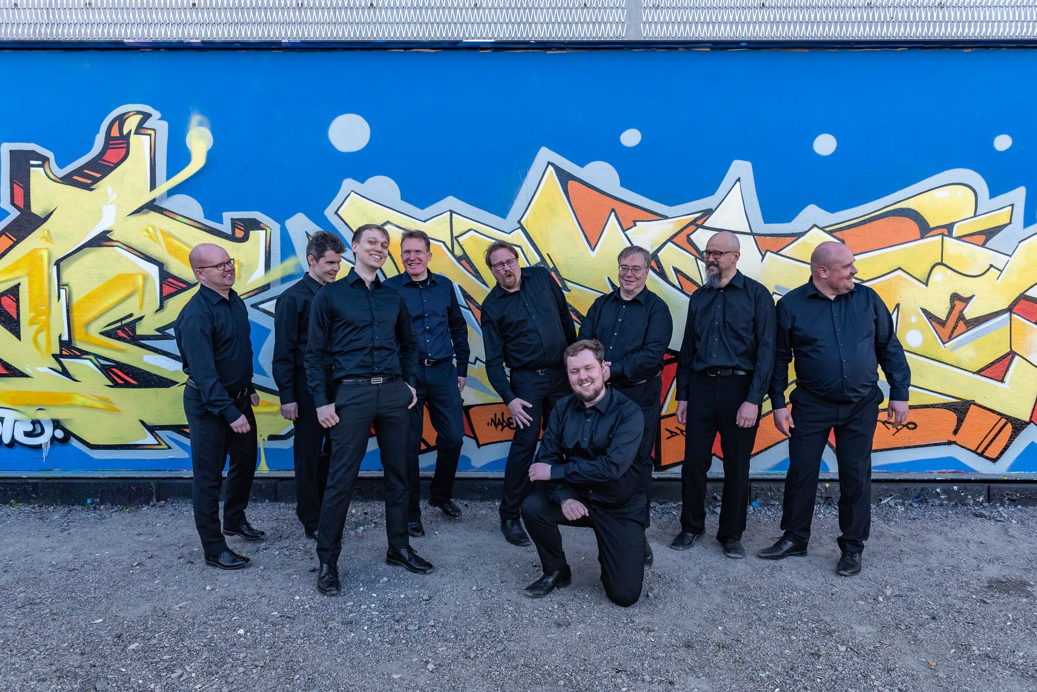 Mustiinpukeutuneet kuoron mieslaulajat poseeraavat nauraen graffitiseinän edessä