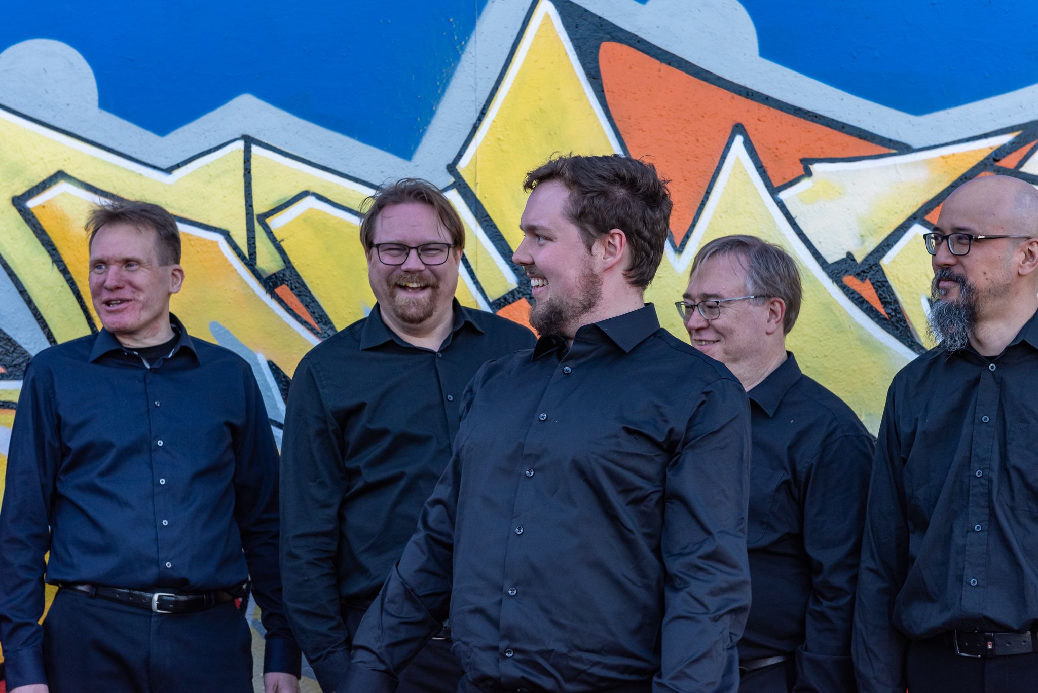 Osa kuoron mieslaulajista nauraa graffitiseinän edessä