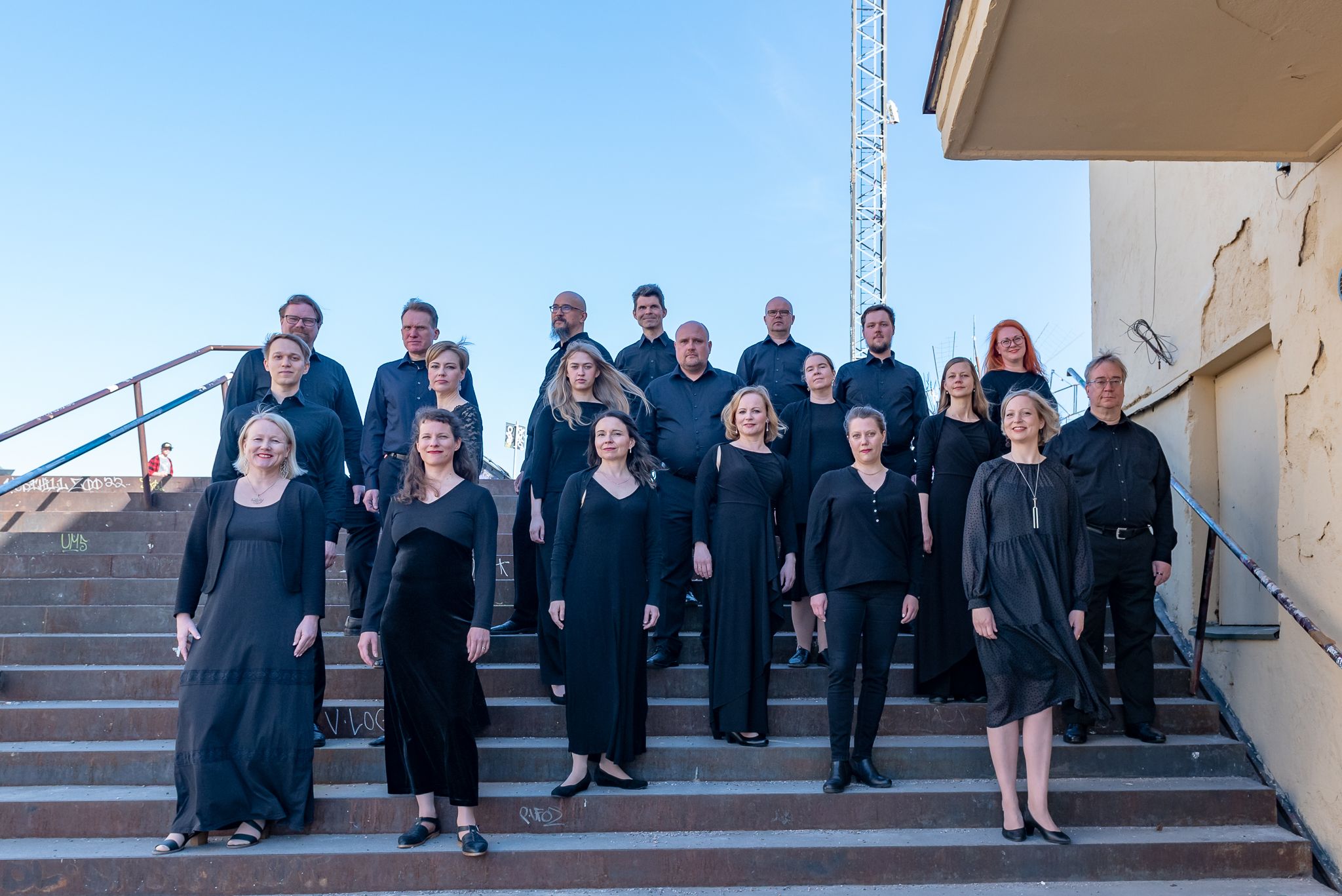 Mustiin pukeutuneet kuorolaiset ryhmäkuvassa portailla pilvetön taivas taustalla